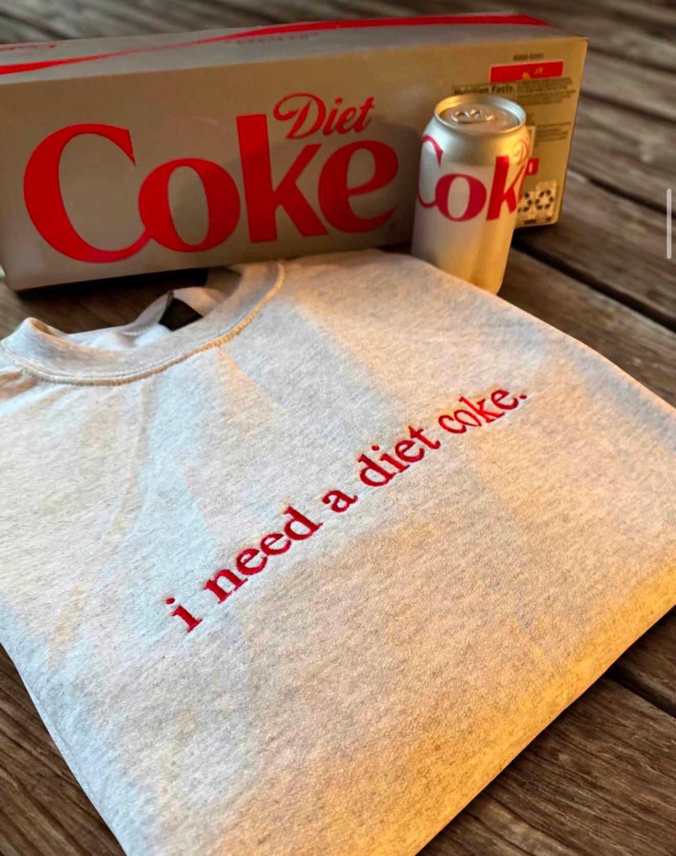 Heather grey tee embroidered in red script reading i need a diet coke, next to a Diet Coke box and can