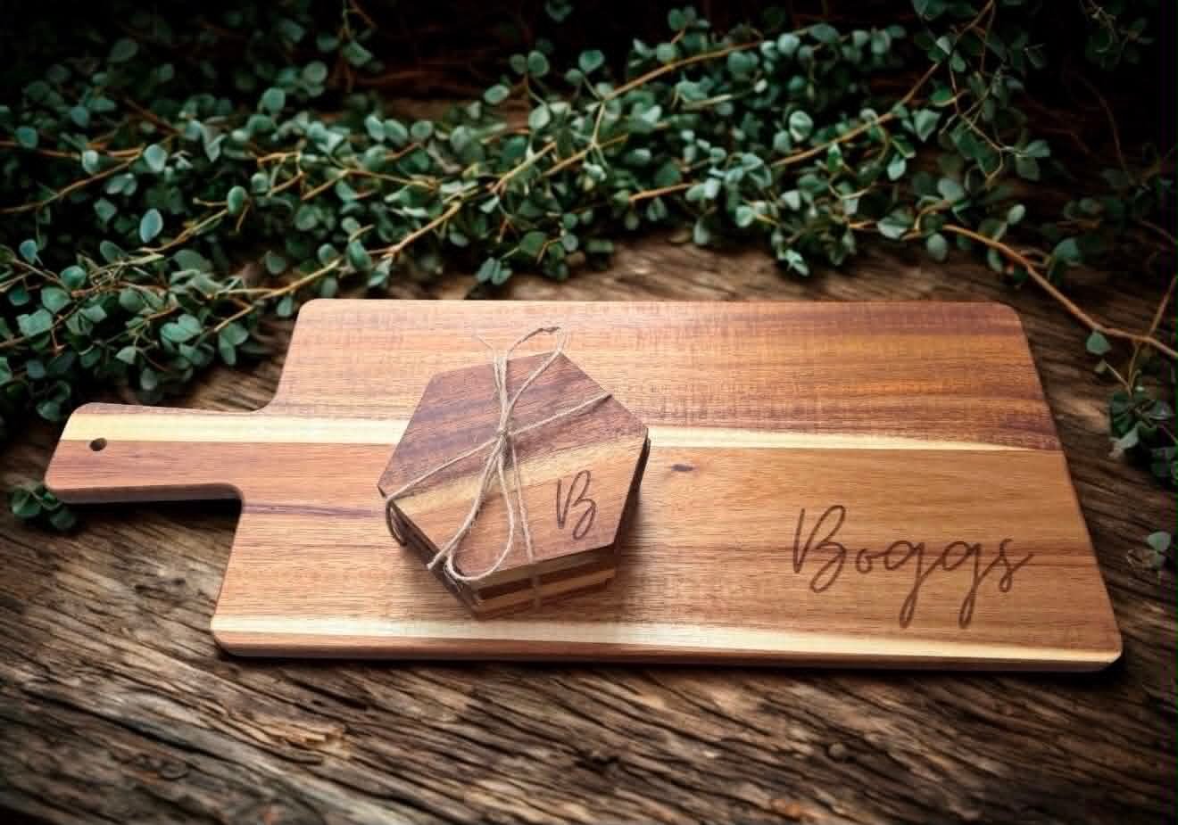 Engraved acacia wood charcuterie board reading Boggs with a bundled hexagon coaster set on top